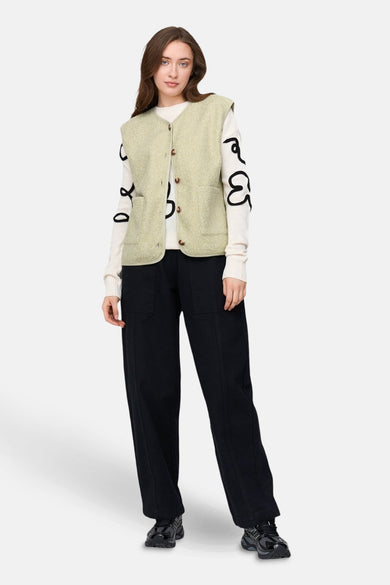 Veste verte sans manches de 24 Colours GMBH, avec des boutons bruns et des poches broiquées, portée avec un pull blanc et un pantalon noir.