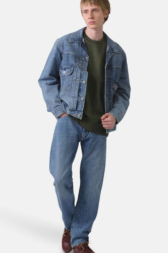 Man met spijkerbroek, denim jack en groene trui staand tegen een witte achtergrond.