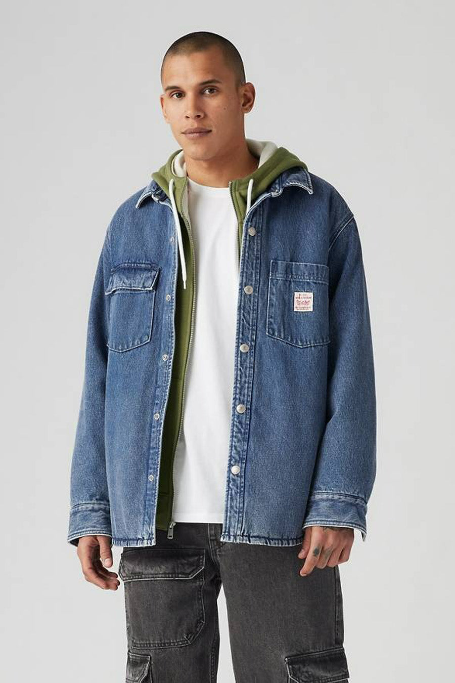 Een man die een blauw denim jack over een witte T-shirt en groene hoodie draagt, met zwarte cargobroeken.
