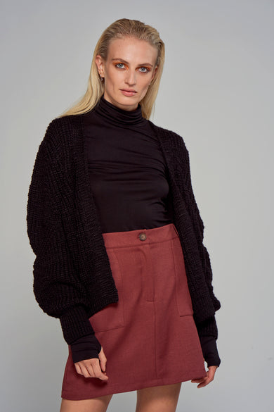 Gilet noir d'Astrid Black Label, avec une texture tissée large, porté sur une veste noire et une jupe rouge.