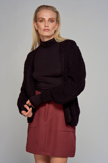 Gilet noir d'Astrid Black Label avec une texture grossière tr tricotée, porté sur un manteau noir et une jupe bordeaux.