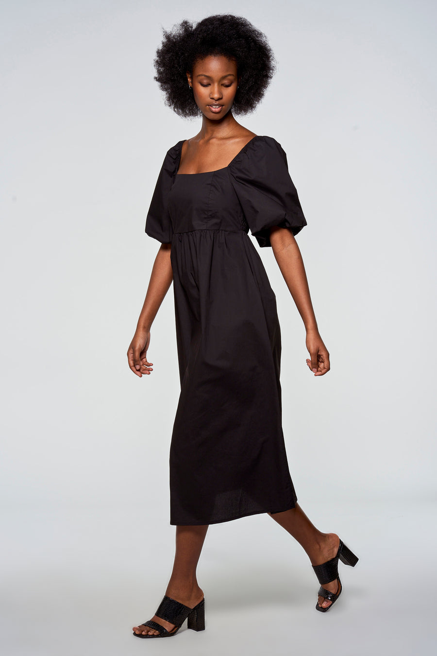 Model in een zwarte katoenen jurk met pofmouwen en zwarte hakken.