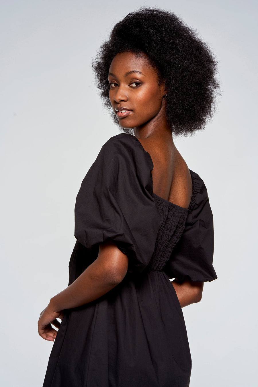 Vrouw met afro in zwarte jurk, over de schouder kijkend, witte achtergrond.
