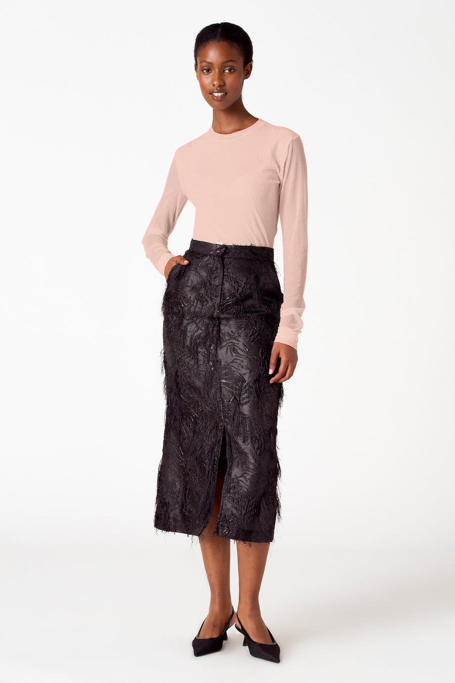 Model in perzikkleurige top en zwarte rok met splitten, staand tegen een witte achtergrond.