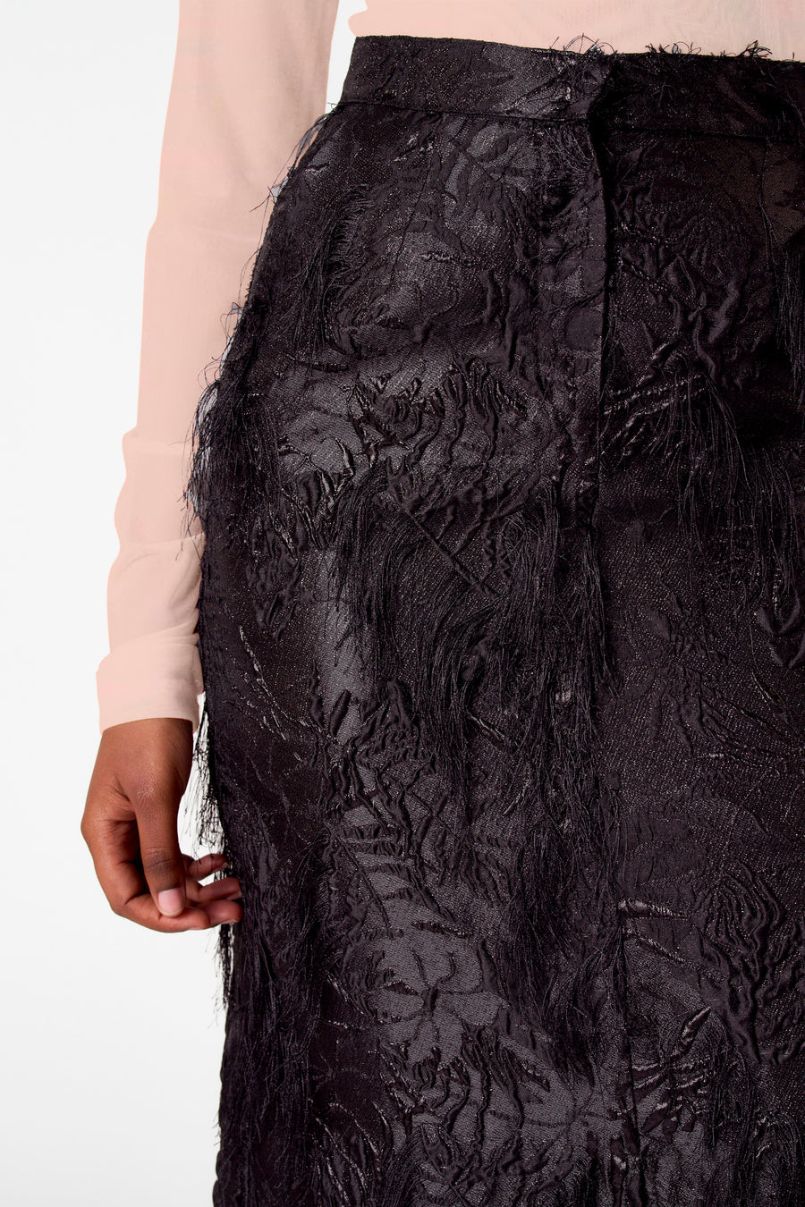 Close-up van een zwarte rok met textuur en lange, gerafelde details, gedragen met een lichtroze blouse.