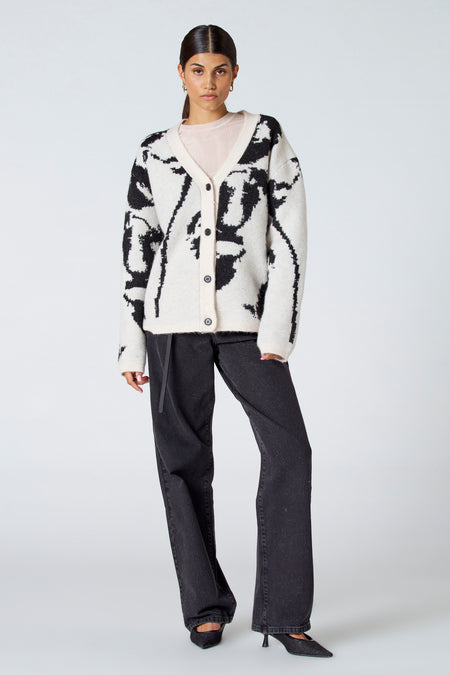 Gilet écru d'Astrid Black Label avec un motif noir, porté sur une chemise rose clair et des jeans à bottes noirs.