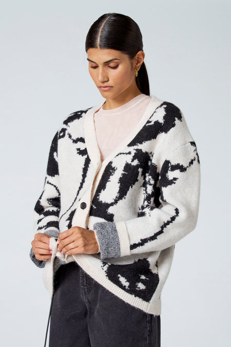 Gilet écru d'Astrid Black Label, avec un dessin noir et des manches longues, porté sur une chemise rose et un pantalon noir.