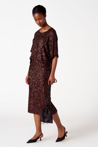 Vrouw in bruine paillettenjurk met zwarte handtas en slingback hakken.
