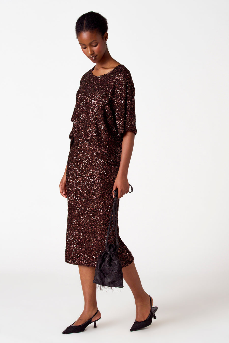 Vrouw in bruine paillettenjurk met zwarte handtas en slingback hakken.
