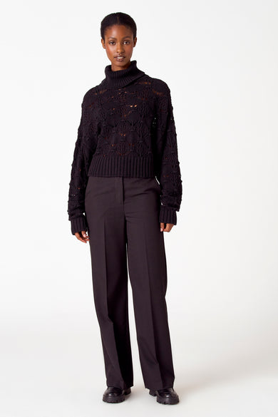 Pull noir à col montant d'Astrid Black Label, avec un motif ajouré et des bordures côtelées, associé à un pantalon et des chaussures.