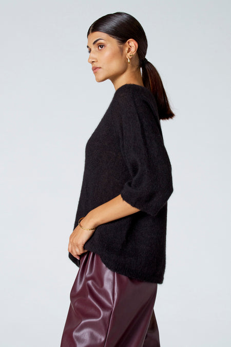 Zwarte trui met ronde hals van Astrid Black Label, gedragen met bordeauxkleurige leren broek en gouden armband, in zij-aanzicht.
