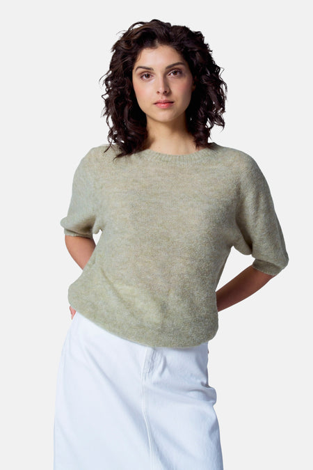 Pull vert avec col rond d'Astrid Black Label, à manches courtes, porté avec un pantalon blanc.