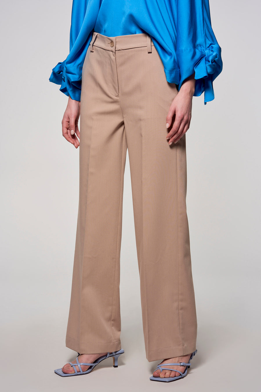 Een persoon die een blauwe blouse en beige, wijde pantalon draagt, met blauwe hakken.