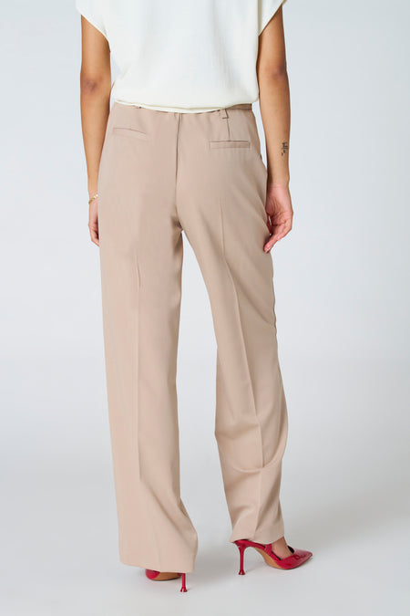 Pantalon beige Astrid Black Label, avec longues poches, poches arrière et associé à des talons rouges.