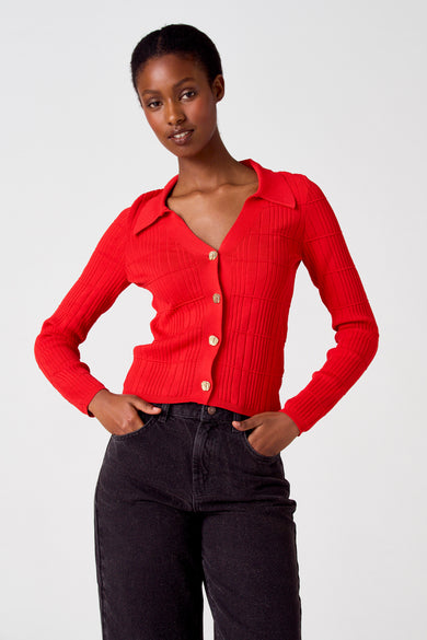 Gilet rouge d'Astrid Black Label, avec des manches longues et des boutons dorés, associé à un jean noir.