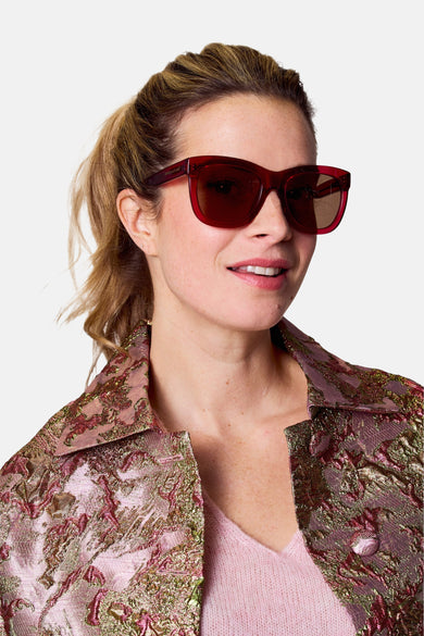 Lunettes de soleil roses d'Astrid Black Label, portées par une femme avec un manteau rose à motif brocart.