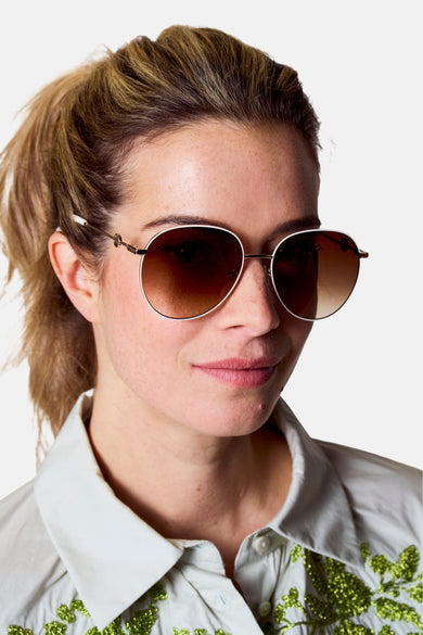 Lunettes de soleil blanches d'Astrid Black Label, avec un cadre doré et des verres marrons, portées par une femme en chemise verte claire.