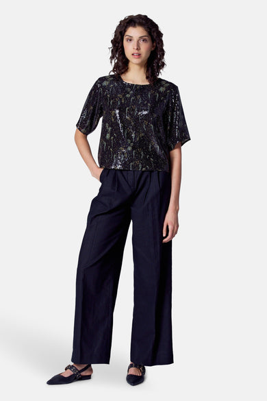 Vrouw met zwarte glinsterende top en donkerblauwe broek, staand op een witte achtergrond.