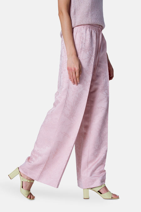 Pantalon rose d'Astrid Black Label, avec un motif floral tissé et des jambes larges, associé à des sandales jaunes claires.