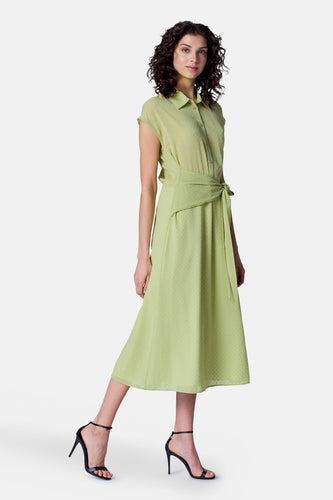 Vrouw met krullend haar draagt groene blousejurk en zwarte hakken, staand op een witte achtergrond.