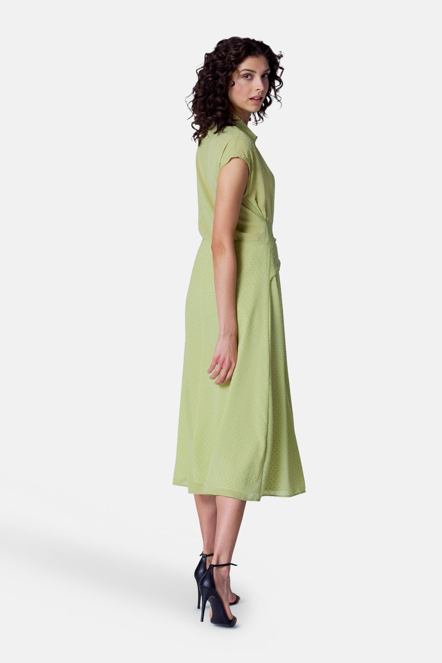 Vrouw in groene jurk met hakken, staand op witte achtergrond.