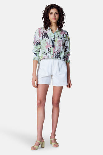 Blouse multicolore à longues manches d'Astrid Black Label, avec un imprimé de fleurs et portée avec des shorts blancs et des sandales jaunes.