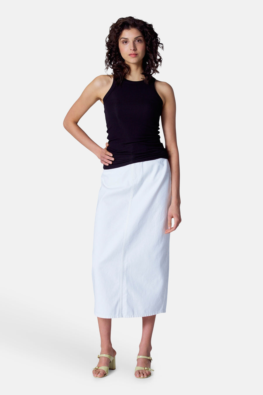 Vrouw in zwarte top en witte rok met groene sandalen staand op witte achtergrond.
