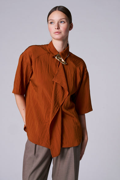Blouse brune Astrid Black Label avec des manches courtes, une ligne de col boutonnée avec un accessoire doré et un pantalon taupe.