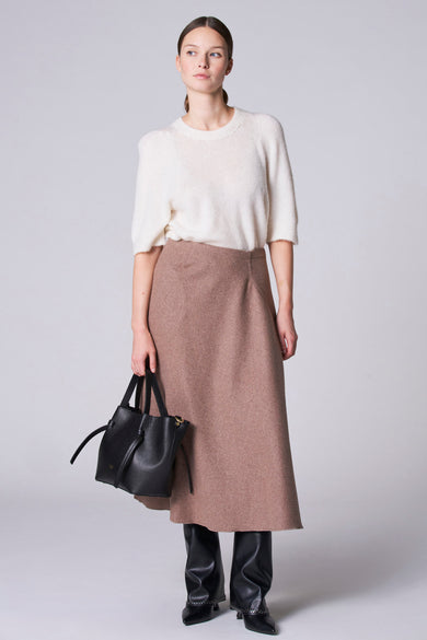 Long jupe taupe d'Astrid Black Label, avec une ligne A fluide et associée à un pull blanc et des bottes noires.