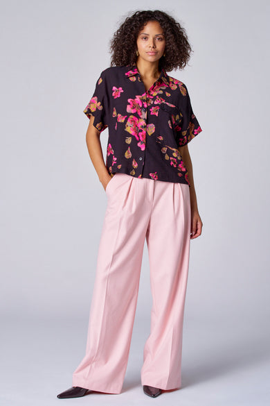 Blouse noire à imprimé de fleurs d'Astrid Black Label, portée avec un pantalon rose large et des chaussures foncées.