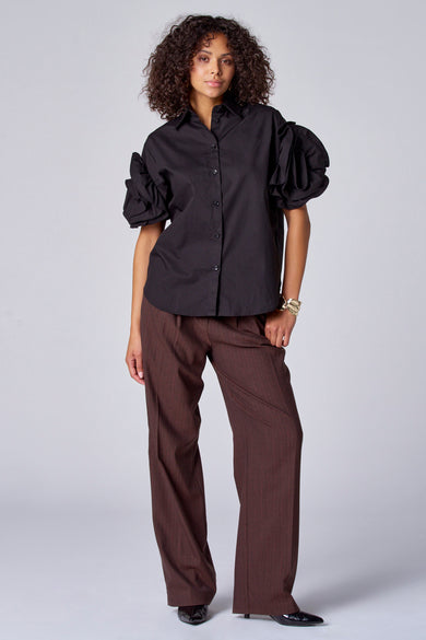 Blouse noire à manches courtes d'Astrid Black Label, avec des manches bouffantes et portée avec un pantalon marron et un bracelet doré.