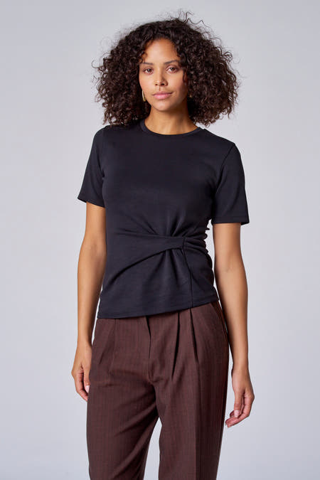 T-shirt noir à manches courtes d'Astrid Black Label, avec un détail de nœud à l'avant, associé à un pantalon marron.