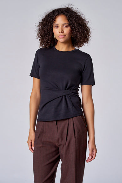 T-shirt noir à manches courtes d'Astrid Black Label, avec un détail de nœud à l'avant, associé à un pantalon marron.