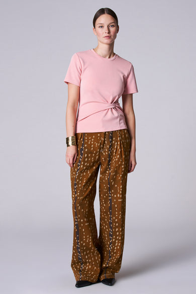 T-shirt rose à manches courtes d'Astrid Black Label, avec un détail de nœud sur le côté, associé à des pantalons marron à carreaux et à un bracelet doré.