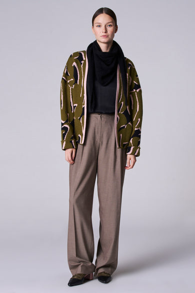 Manteau court vert de Astrid Black Label, avec un motif en noir et rose, porté avec une écharpe noire et des pantalons marron.