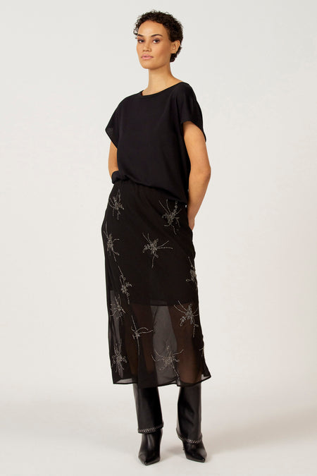 Vrouw in zwarte top en rok met zilveren spinnen, zwarte laarzen, witte achtergrond.