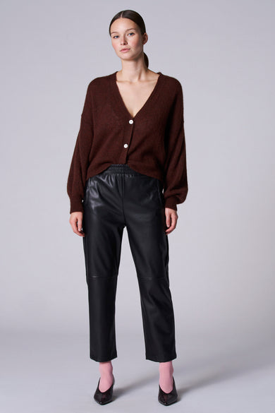 Gilet brun d'Astrid Black Label, avec des manches longues et des boutons blancs, porté avec un pantalon de cuir noir.