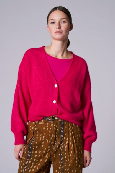 Gilet rose d'Astrid Black Label, avec des manches longues et une fermeture à boutons, associé à un pantalon marron à carreaux.
