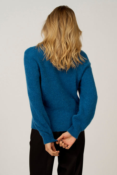 Vrouw met lange blonde golven in een blauwe trui, zwarte broek, achteraanzicht.
