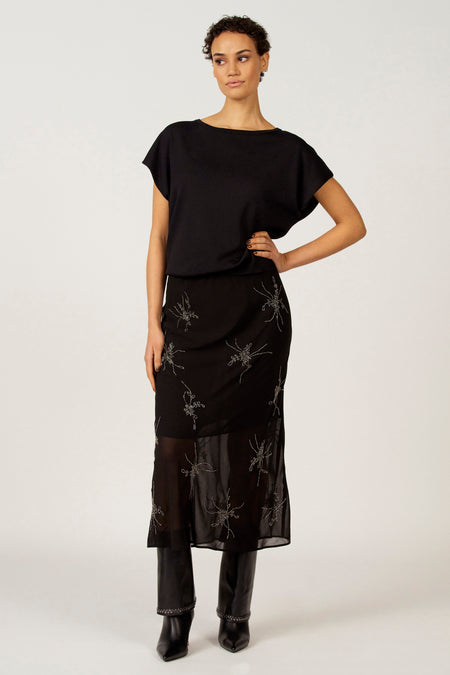 Vrouw in zwarte top en rok met broderie en zwarte laarzen, staand voor witte achtergrond.
