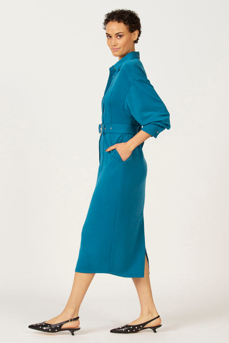 Vrouw in blauwe overhemdjurk met riem en zwarte hakken.