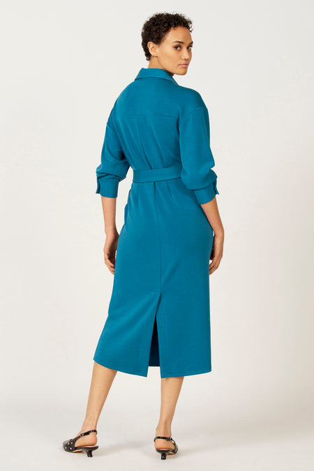 Vrouw in een blauwe jurk met riem, gezien van achteren, met zwarte schoenen.