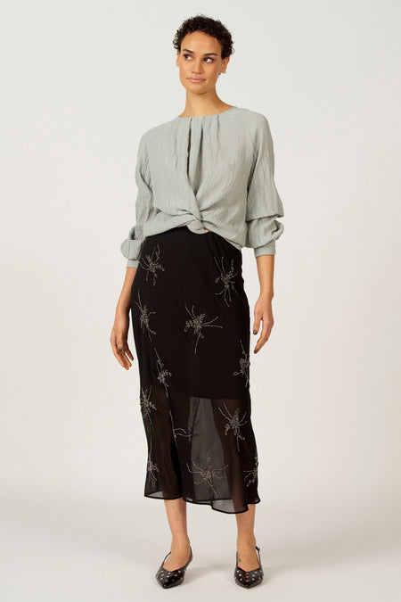 Model draagt een lichtblauwe blouse en een zwarte rok met zilveren accenten.