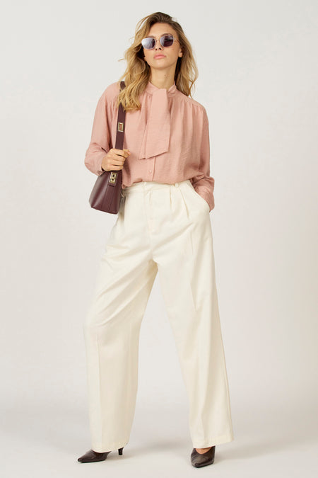 Vrouw in roze blouse, witte broek en bruine handtas.