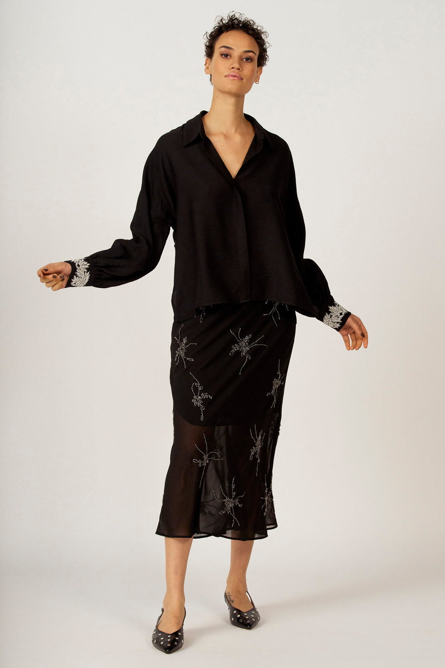 Chemise noire à longues manches d'Astrid Black Label, avec des détails argentés sur les poignets. Portée avec une jupe et des chaussures.