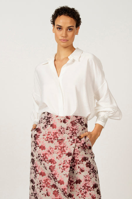 Vrouw in witte blouse en bloemenrok met handen in zakken.