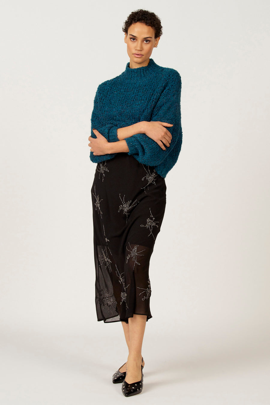 Vrouw in blauwe trui en zwarte rok met zilveren accenten, armen gekruist.