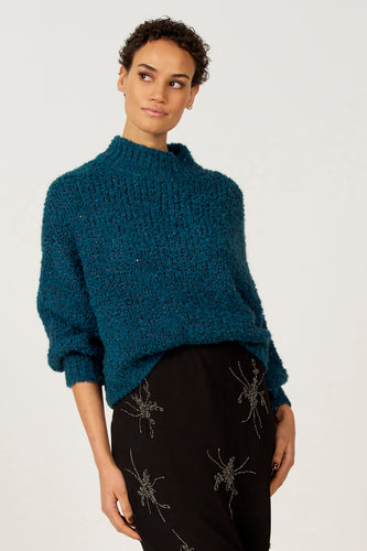 Vrouw met blauwe trui en zwarte rok met versiering, staand voor een witte achtergrond.
