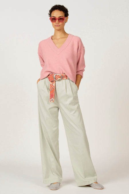 Vrouw in roze trui en groene broek, met riem en roze zonnebril.