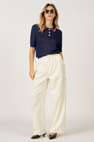 Vrouw in blauwe top met rode kraag, witte broek en zwarte schoenen.